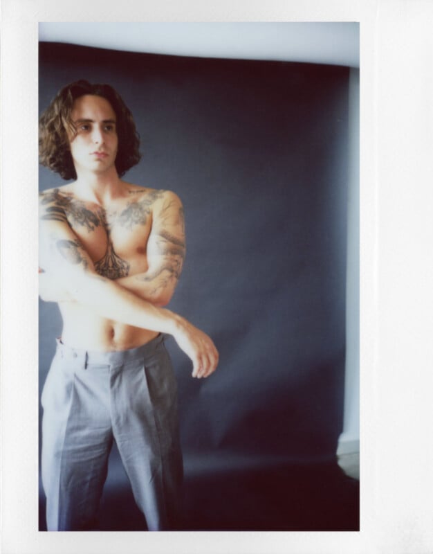 Person with long hair and tattoos stands shirtless against a dark background, wearing gray pants. They have their arms crossed.