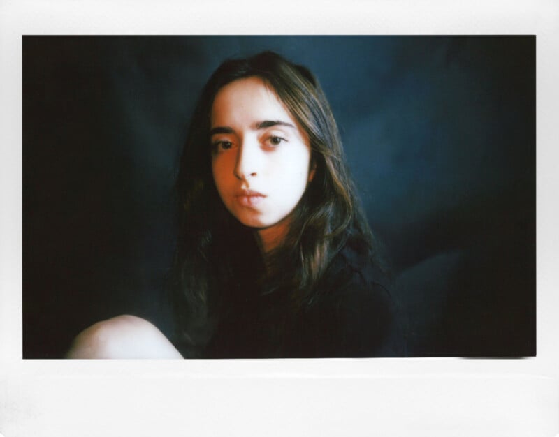 A person with long hair sits against a dark background, illuminated by soft lighting. They have a calm, contemplative expression and are wearing a dark top.