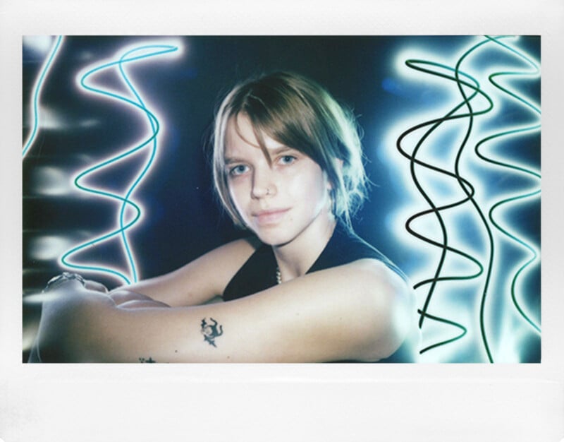 Person with short hair and a nose ring poses with arms crossed. They have a tattoo on their arm and are surrounded by abstract neon light patterns. The background is dark, highlighting the person and the glowing lights.