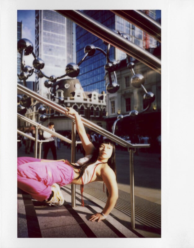 A person in bright pink pants and running shoes poses on an outdoor stair railing, surrounded by modern architecture with glass buildings and sculptures. The sun casts dramatic shadows across the scene.