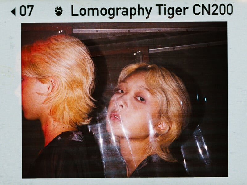 A stylized photo of two people with light blonde hair against a dark background. The person on the right looks forward, while the person on the left faces away. The image has a Lomography Tiger CN200 film border and a soft red hue.