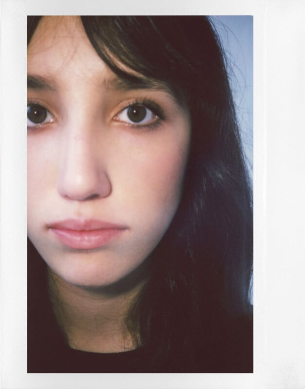 Close-up portrait of a person with long dark hair and brown eyes, looking directly at the camera. The background is softly blurred, emphasizing the subject's facial features.