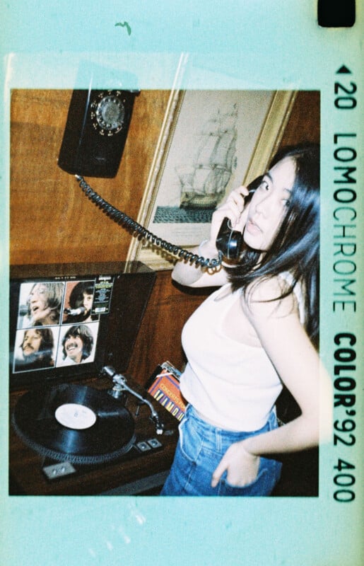 A woman in a white tank top and jeans uses a rotary phone. Nearby, a vinyl record player with album covers on the wall adds a vintage touch. The scene is framed in a retro style with "LomoChrome Color '92" branding.