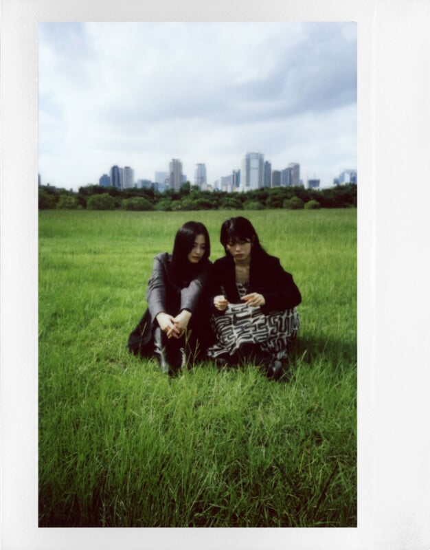 Two people are sitting on grass in an open field with a city skyline in the background. They appear to be looking at a phone together, surrounded by green grass under a cloudy sky.