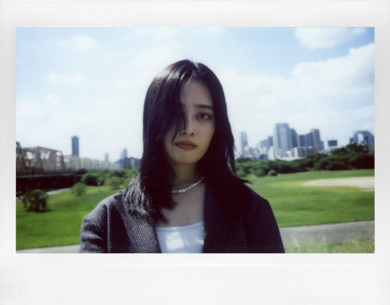 A woman with long black hair stands in a grassy area with a city skyline in the background. She is wearing a dark jacket and a pearl necklace. The sky is blue with some clouds, and a bridge is visible to the left.