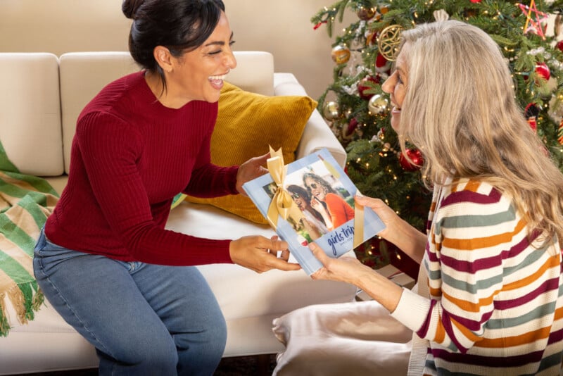 Two women sitting on a couch near a decorated Christmas tree exchange gifts with joyful expressions. One woman holds a wrapped book with a ribbon, featuring a photo of two people on the cover.