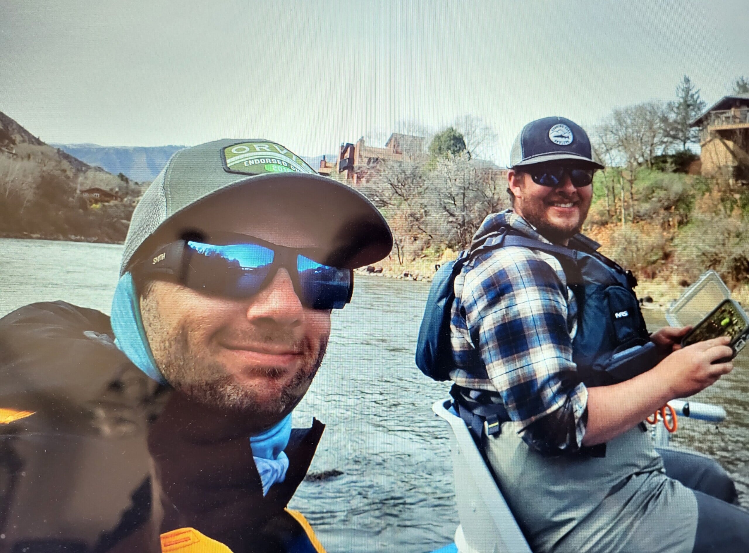 Two people are sitting in a boat on a river. They are wearing hats and sunglasses. The person on the left is taking the photo, while the person on the right is smiling. Trees and houses are visible in the background.