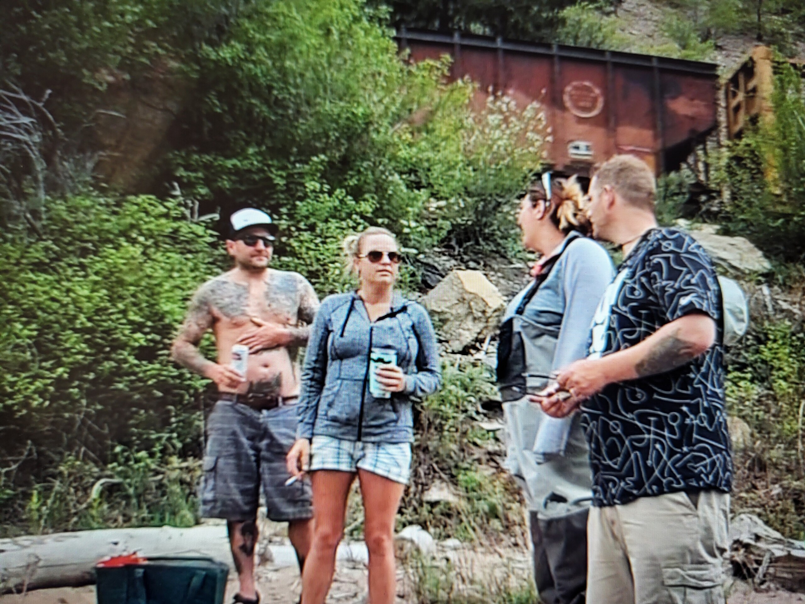 Four people stand outside near greenery and a railroad car. One person is shirtless with tattoos, another wears sunglasses and holds a can, while the others are in casual clothing. They're engaged in conversation in a wooded area.