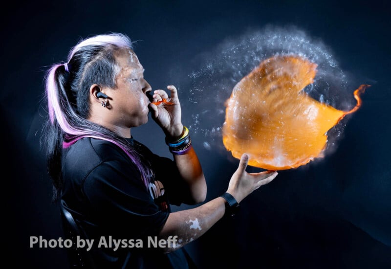 A person with long, two-toned hair blows a balloon, which bursts in an explosion of water, captured mid-splash against a dark background.