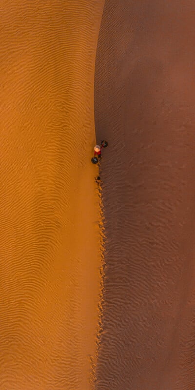 Aerial view of a person in an orange outfit walking on a sand dune, leaving footprints behind. The sand displays two distinct textures, with the left half smooth and the right half ridged, creating a striking contrast. The lone figure casts a shadow on the surface.