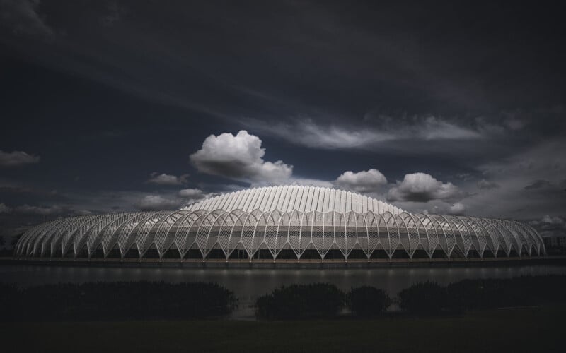 A futuristic building with a sleek, geometric design and white arches stands against a dramatic sky with fluffy clouds. The structure is reflected in calm water in the foreground.