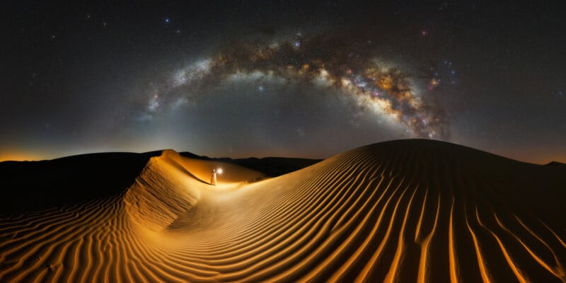 A lone person stands on a dune with a lantern in hand in a vast desert at night. The sky above is illuminated by the Milky Way, creating a striking arc of stars over the scene. Shadows and light highlight the ripples in the sand.