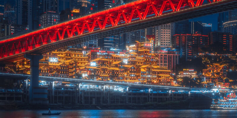 A vibrant nighttime cityscape featuring a brightly lit, traditional-style building complex under a striking red bridge. The glowing structures, set against modern high-rises, reflect on the calm water below, where a single small boat can be seen.