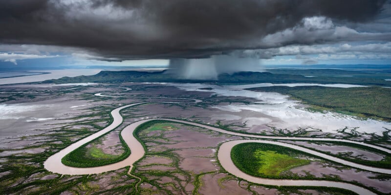 Aerial view of a vast landscape with a meandering river flowing through green vegetation and muddy terrain. Dark, ominous clouds overshadow the area, with heavy rain pouring in the distance, creating a dramatic contrast between the light and dark sky.