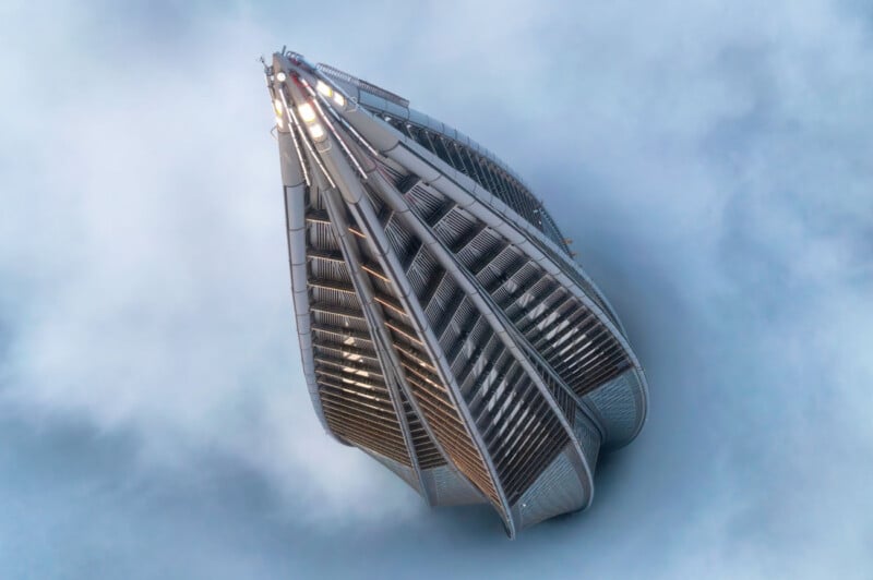 Aerial view of a modern, futuristic skyscraper emerging through thick clouds. The building features a sleek, curved design with multiple tiered, metallic facades converging to a pointed top, creating a shape reminiscent of a ship's bow cutting through the fog.