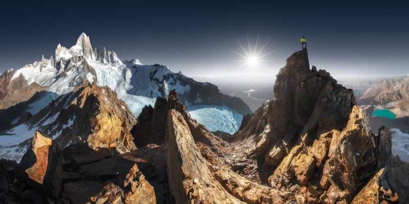 A hiker in a yellow jacket stands on top of a rocky peak under a clear, dark sky. The sun shines brightly over a expansive, rugged mountain landscape with snow-capped peaks and a glacier visible in the distance.