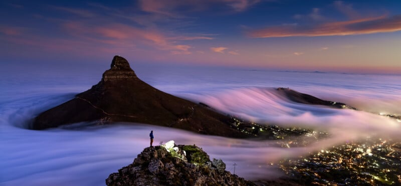 A lone hiker stands on a rocky peak overlooking a breathtaking mountain landscape at sunset. A sea of clouds cascades over the hills and valleys, with a prominent pointed peak rising above the clouds. The sky transitions from deep blue to soft pink hues.