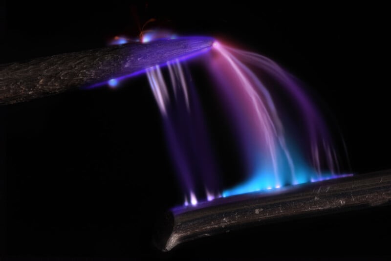 A vivid electrical arc discharges between two metal rods against a dark background. The colorful display features streaks of blue, purple, and white light, creating a dynamic and energetic visual effect.