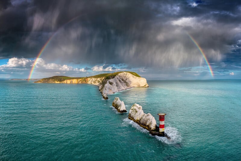 A scenic view of a lighthouse on a rocky coastline under a dramatic sky. A vivid rainbow arches over the scene, contrasting against dark storm clouds. The lighthouse, with red and white stripes, stands near the edge of turquoise waters.