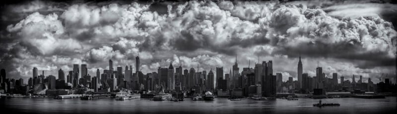 A panoramic black-and-white photo of a city skyline featuring numerous high-rise buildings. The cloudy sky looms dramatically above the skyscrapers, creating a striking contrast. The waterfront in the foreground includes docks and low-rise structures.