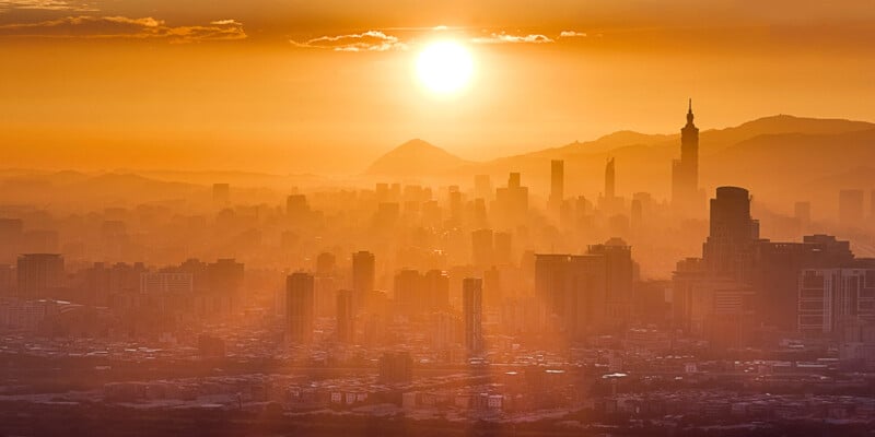 A city skyline is bathed in a warm orange glow as the sun sets in the background. High-rise buildings and towers rise prominently, with mountains shadowed behind them. Sun rays pierce through the atmosphere, casting a serene and golden ambiance over the scene.