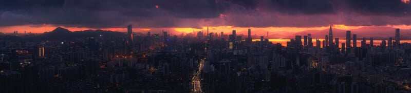 A panoramic view of a cityscape at twilight. Skyscrapers and buildings are silhouetted against a dramatic sky with dark clouds and vibrant shades of orange, pink, and purple from the setting sun. The city lights begin to illuminate the urban landscape.