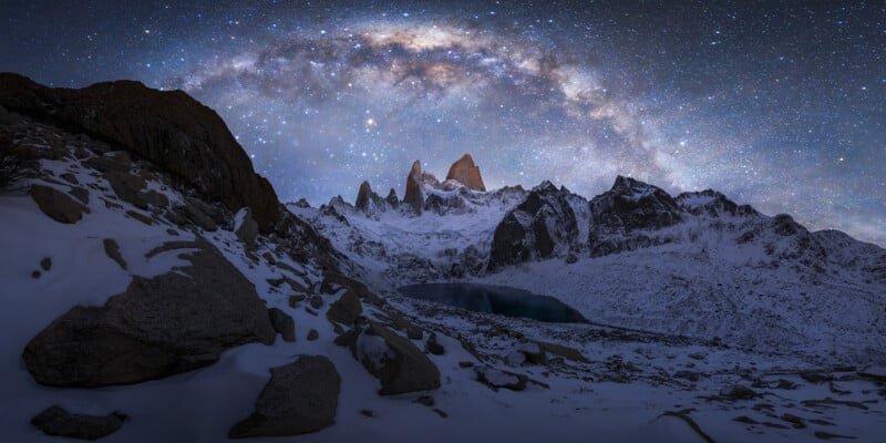 A breathtaking night view of a snow-covered mountain range with jagged peaks under a star-filled sky. The Milky Way is visible, arching majestically over the peaks. Snow and rocky terrain are illuminated by starlight, adding to the ethereal beauty of the scene.