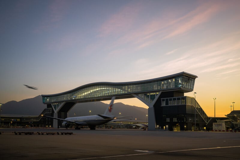 A modern airport terminal at dusk, featuring a sleek, wave-shaped structure. A plane is parked at the terminal, while another is taking off against a backdrop of mountains and a colorful sunset sky.