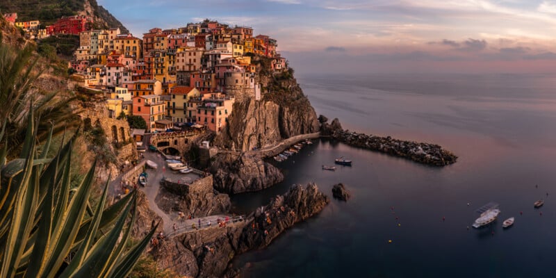 Panoramic view of Manarola, one of the five villages of Cinque Terre in Italy. Colorful houses are perched on a rocky cliff overlooking the serene sea at sunset. Boats are moored in the small harbor below, and the sky is painted with soft hues.