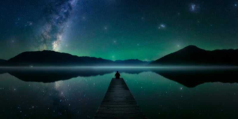 A person sits at the end of a wooden dock extending over a calm lake, gazing at a breathtaking night sky filled with stars and the Milky Way. Majestic mountains are silhouetted against the celestial backdrop.