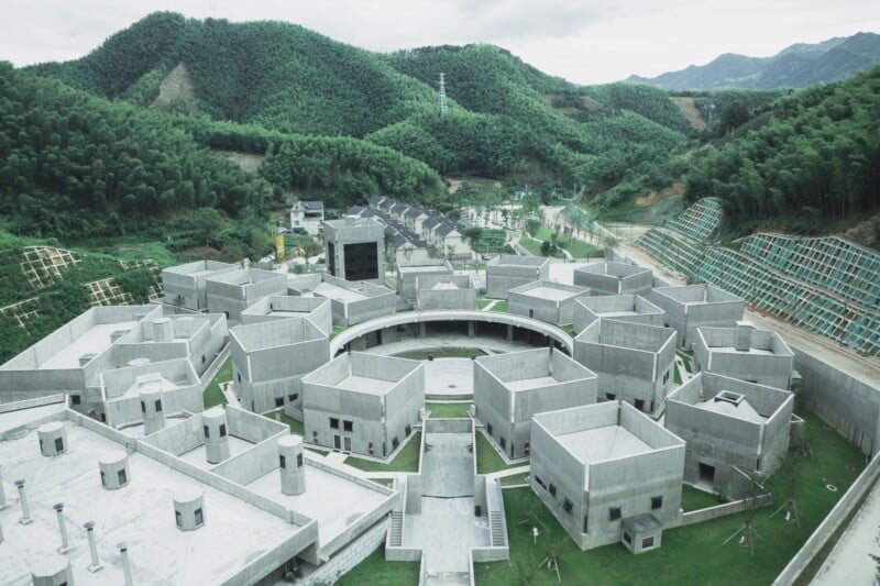 Aerial view of a contemporary concrete building complex with geometric structures and a central courtyard, surrounded by lush green hills and trees. The background features additional buildings and mountainous terrain.