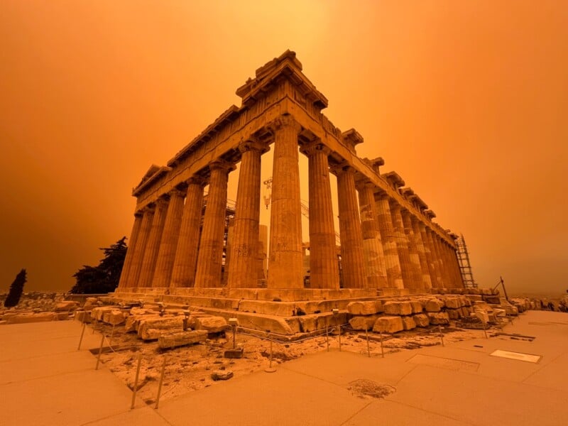 The image shows the Parthenon on the Acropolis of Athens under an orange sky, giving the scene a dramatic, warm hue. The ancient temple's columns and stone structure are prominent in the foreground.