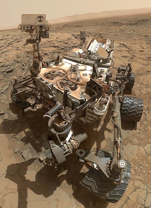 A large, complex rover with six wheels and a tall mast camera stands on a rocky, reddish surface, displaying various scientific instruments and panels. The landscape has a barren, dusty appearance.