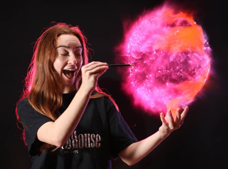 A person with long hair and a black T-shirt is excitedly using a stick to hold a glowing, colorful orb with pink and orange hues against a dark background.