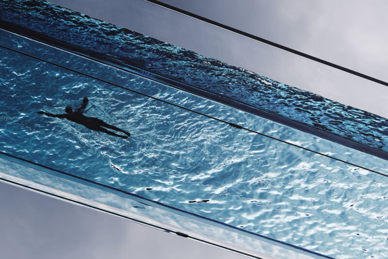 A person swimming in a clear, elevated glass pool against the sky, with water rippling around them. The perspective from below emphasizes the pool's transparency and the swimmer's silhouette.