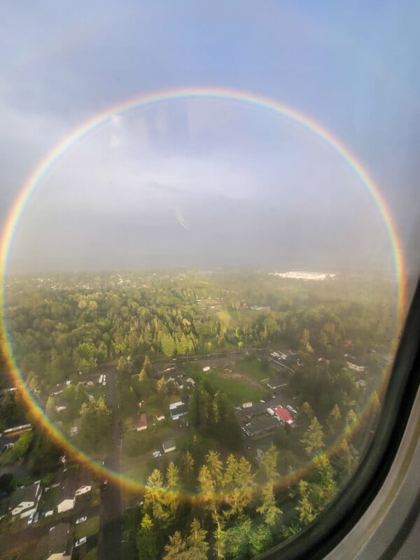 Aerial view from an airplane window showing a complete circular rainbow over a landscape with lush green trees and scattered houses. The sky is partly cloudy with soft sunlight illuminating the scene.