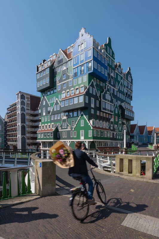 A cyclist rides past a uniquely designed building in Zaandam, Netherlands. The structure features stacked, colorful houses typical of the region's architectural style, with blue skies in the background.