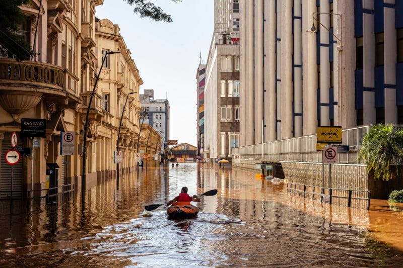 A person in a red shirt paddles a kayak down a flooded street between tall buildings. The water reflects the architecture, and there are street signs visible. The scene suggests an urban area experiencing severe flooding.