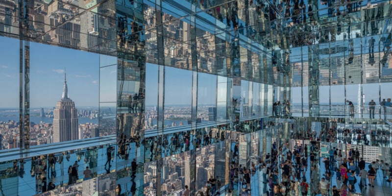 A large crowd of people is gathered in a modern observatory with floor-to-ceiling glass walls and mirrors, creating intricate reflections. The observatory overlooks a cityscape, featuring a prominent skyline with the Empire State Building.