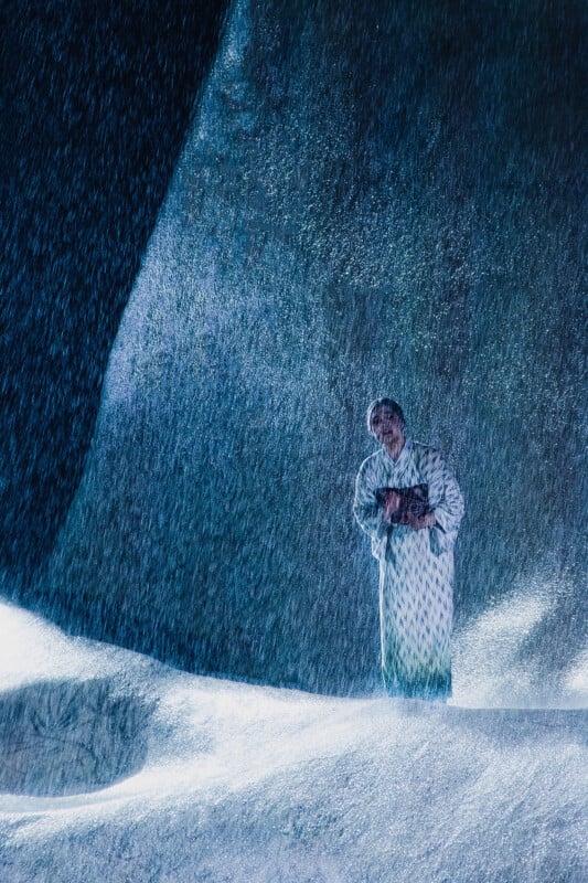 A person in a white coat stands alone holding a book, surrounded by large, dark icy formations. Snow falls heavily around them, creating a dramatic wintry scene.