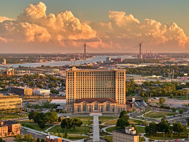 Aerial view of a large, historic building surrounded by greenery in an urban area. Behind it, a river winds through the cityscape, with bridges and industrial structures. The sky is filled with dramatic, fluffy clouds illuminated by golden light.