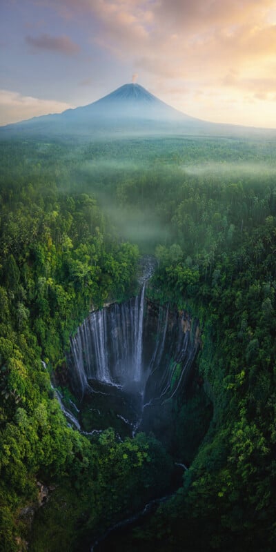 An aerial view of a breathtaking landscape featuring a lush green forest surrounding a deep crater with a waterfall cascading down its walls. In the background, a majestic, snow-capped mountain stands tall under a colorful, partly cloudy sky at sunrise or sunset.