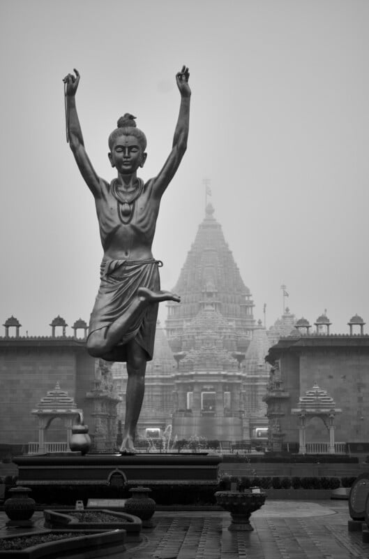 A grayscale image of a tall statue depicting a robed figure with arms raised, standing on one leg. In the background, a large ornate temple with intricate carvings looms under a cloudy sky, surrounded by lower walls and smaller structures.