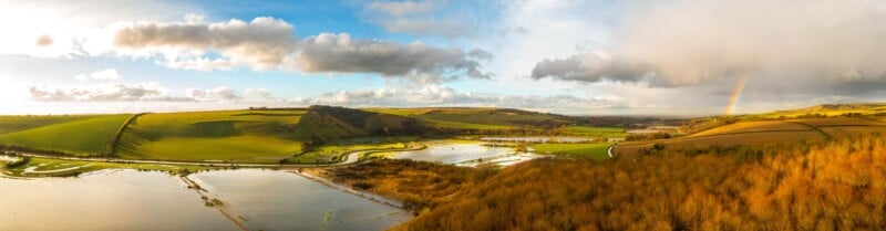 A panoramic view of a rural landscape with rolling green hills and fields under a partly cloudy sky. A rainbow is visible on the right, above patches of water reflecting the sky, bordered by leafless trees in the foreground.