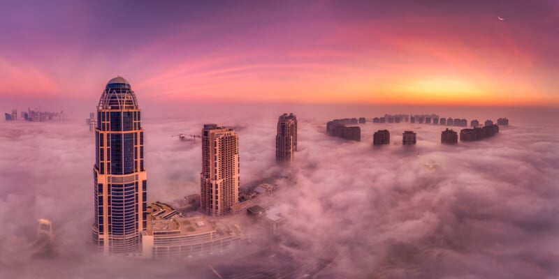 A breathtaking aerial view of a city skyline enveloped in dense fog during sunrise. Tall skyscrapers and residential high-rises pierce through the mist. The sky is painted in vibrant hues of orange, pink, and purple, with a faint crescent moon visible.