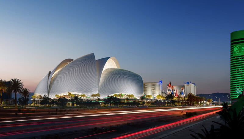 Futuristic architecture illuminated at dusk with multiple white, shell-like structures. A road in the foreground with light trails from moving vehicles. Buildings with colorful lights are visible in the background against a clear sky.