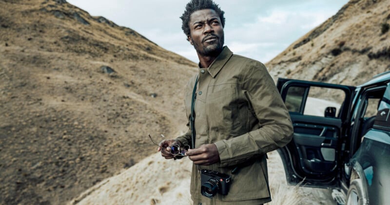 A man in a green jacket stands near an open car door on a mountainous road. He's holding sunglasses and has a camera around his neck. The landscape is rocky and dry, with cloudy skies above.