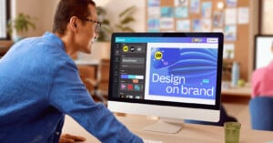 A person in a blue shirt uses design software on a desktop computer. The screen displays a graphic with the text "Design on brand" alongside various editing tools. The workspace has a blurred background with plants and decor.