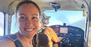 A woman smiles at the camera while sitting in the passenger seat of a small aircraft. The pilot, wearing headphones and a cap, is operating the plane. The cockpit is filled with controls, and the sky is visible through the windows.