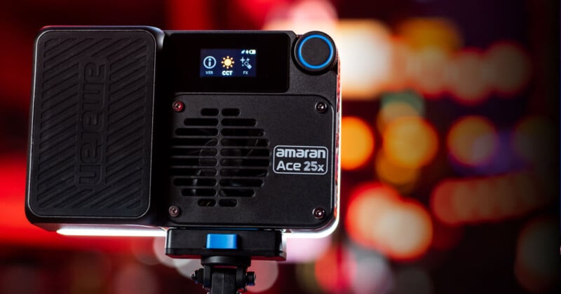 A compact black Amaran Ace 25x light with a digital display and control knobs is mounted on a stand. The background is blurred with colorful bokeh lights in red, orange, and yellow hues.